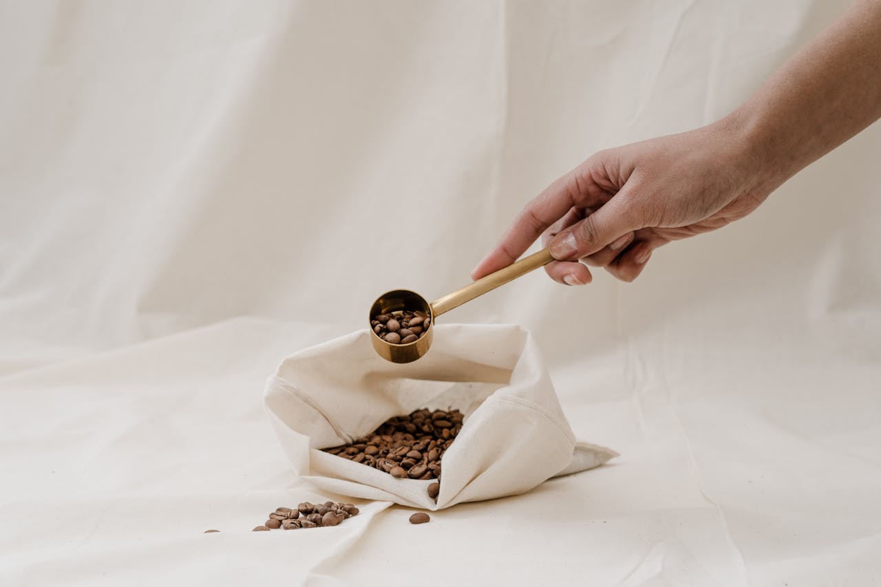 our-journey-02 A hand holding a scoop over a fabric bag filled with coffee beans, highlighting a minimalist aesthetic.